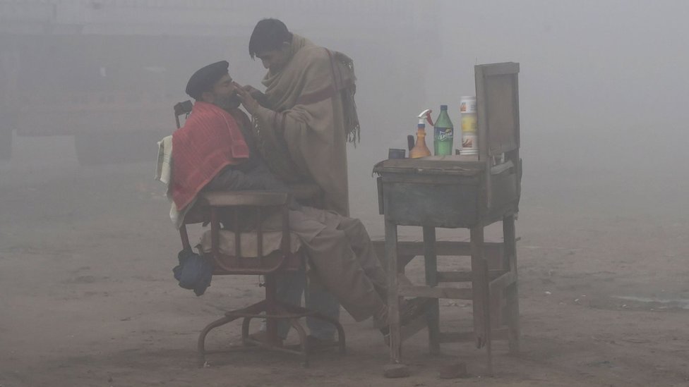 Пакистанский парикмахер бреет клиента на дороге в условиях сильного тумана и смога в Лахоре 24 января 2019 года.