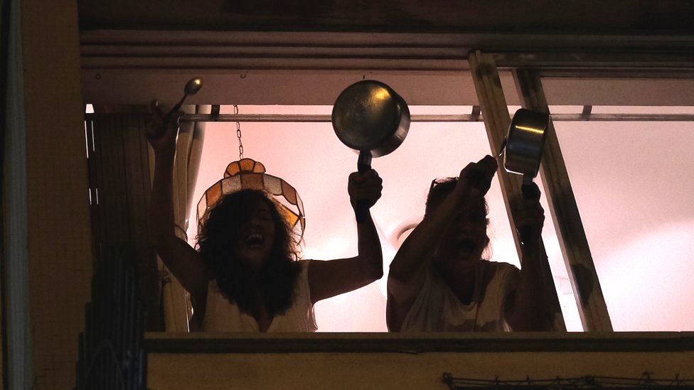 Женщины стучат горшками в окно своей квартиры в Рио-де-Жанейро, протестуя против президента Бразилии Жаира Болсонару по поводу его борьбы с пандемией коронавируса, 19 марта 2020 г.
