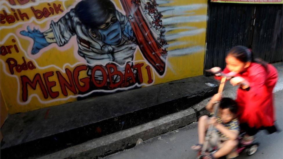 Duas crianças juntas em um patinete na rua, passando ao lado de um grafite com boneco se protegendo do coronavírus
