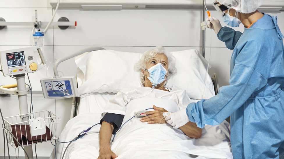 Idosa deitada de máscara em cama de hospital sendo atendida por médico