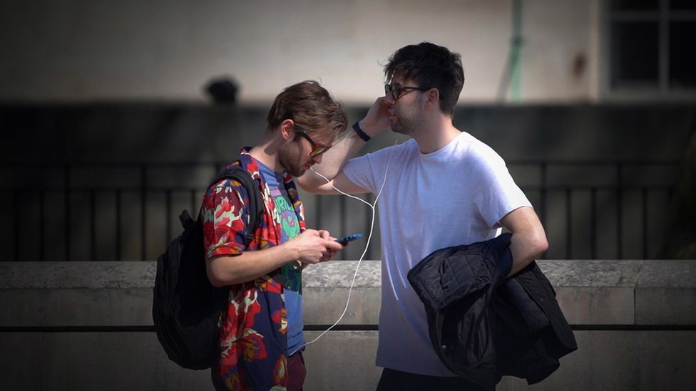 Two men in sunglasses sharing a pair of earphones
