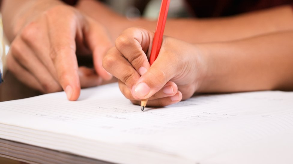 Mão de adulto aponta para caderno em que criança escreve com lápis