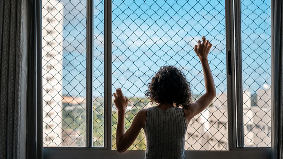 Una mujer de espaldas se agarrra de una red en una ventana mientras mira afuera.