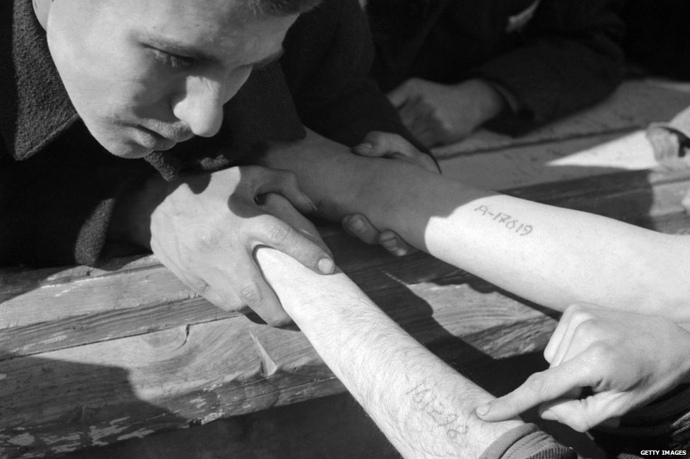 Молодой человек проверяет числа, вытатуированные на руках евреев-польских военнопленных, прибывших из Освенцима, в концлагерь Дахау после его освобождения армией США в конце апреля 1945 года