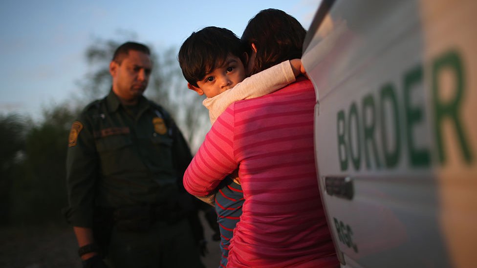 Una mujer sostiene a un niño ante un agente fronterizo