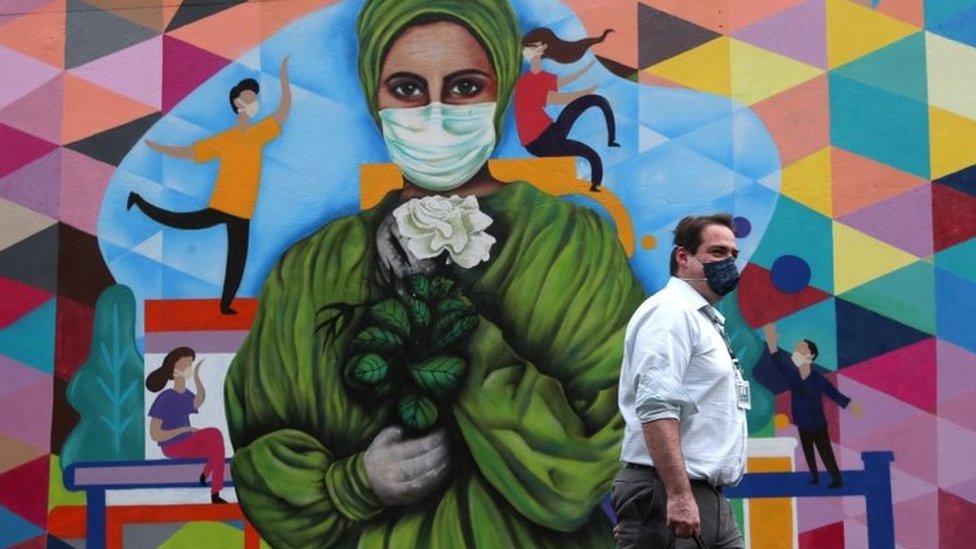 Hombre delante de un mural con un dibujo de mujer con máscara.