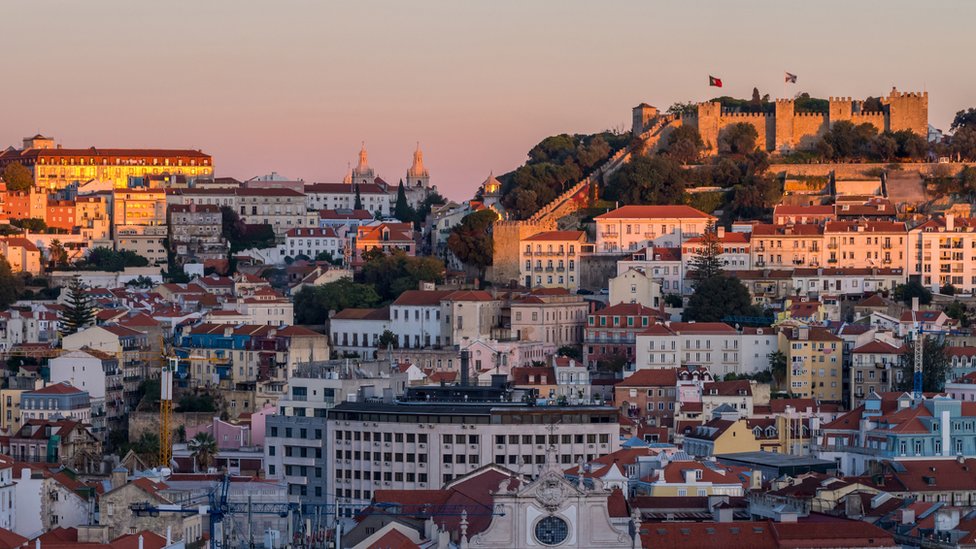 Imagem mostra vista geral do bairro de Alfama, em Lisboa, com vários imóveis, bandeiras do país e o Castelo de São Jorge aparecendo