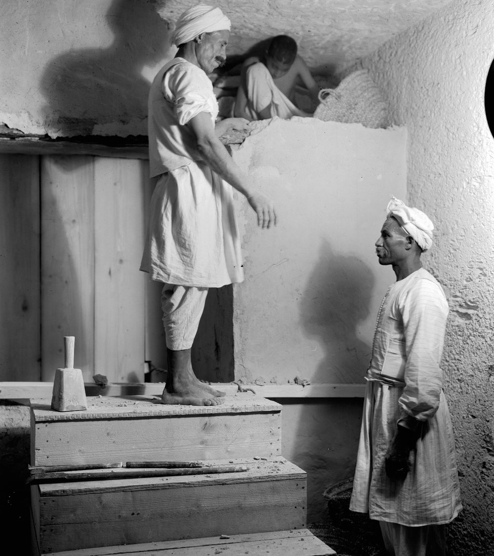 Foto em preto e branco mostra dois trabalhadores egípcios e uma criança desmontando uma parede em um local interno iluminado; um dos trabalhadores está descalço