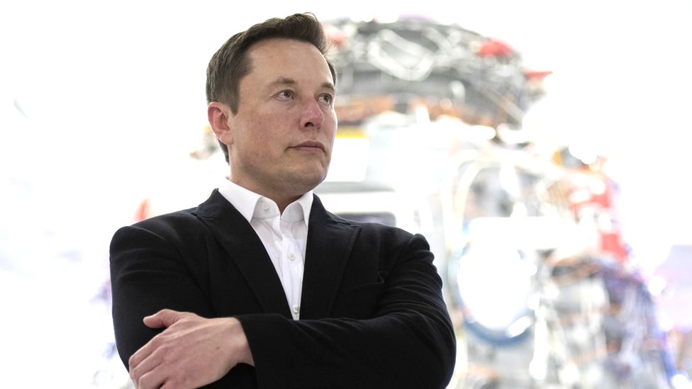 Elon Musk stands in a harshly lit room at SpaceX, in front of a hulking piece of metal and wire