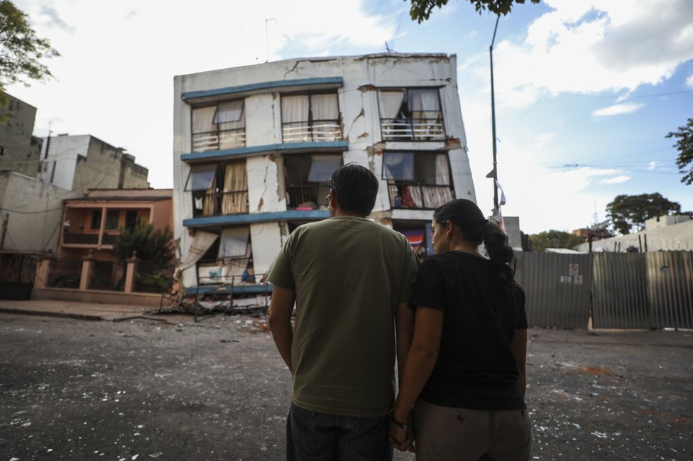 Мужчина и женщина стояли перед поврежденным зданием.То, что раньше было пятиэтажным домом, полностью потеряло два нижних уровня.