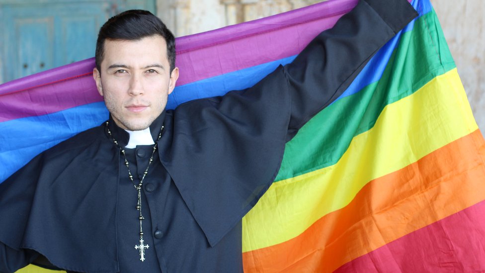 Un sacerdote sostiene una bandera multicolor, símbolo de la comunidad LGBT