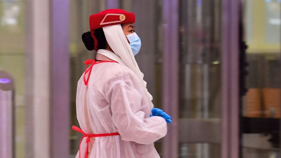 Am Сотрудник авиакомпании (в июле) в защитном снаряжении наблюдает за тем, как туристы проходят медицинский осмотр по прибытии в Терминал 3 в аэропорту Дубая, Объединенные Арабские Эмираты