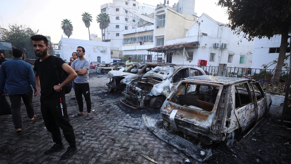 People inspect Al-Ahli hospital with burned out vehicles in the foreground