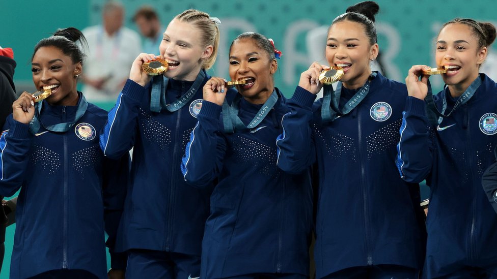 Američki ženski gimnastičarski tim, s leva na desno: Simon Bajls, Džordan Čils, Džejd Keri, Sunisa Li i Hili Rivera drže zlatnu medalju u zubima