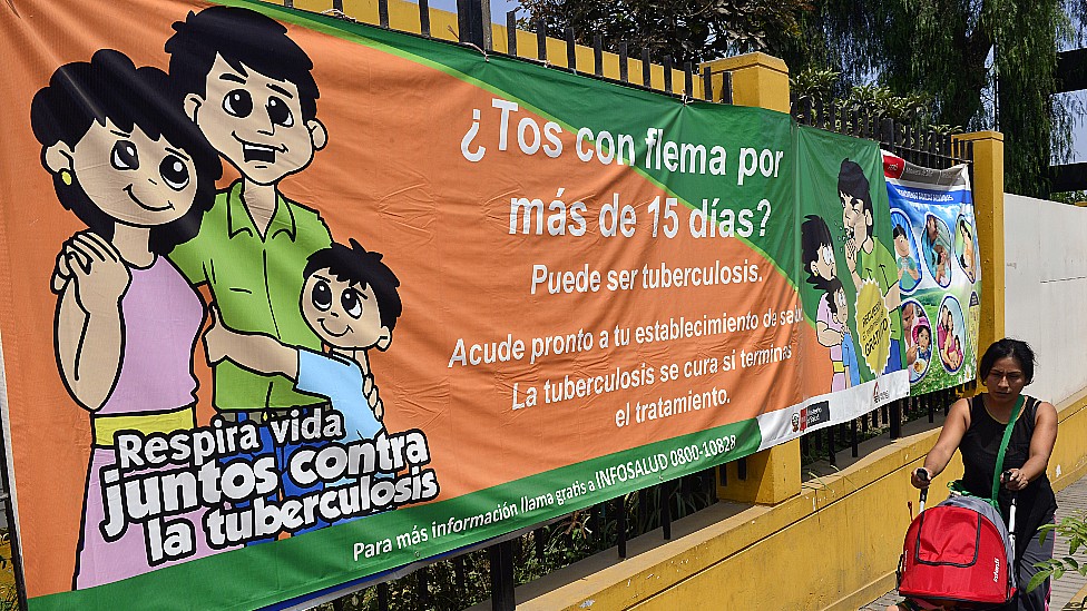 Mujer con su niño en Perú pasando frente a una pancarta que insta a la gente a acudir al médico si han tenido tos con flema por más de 15 días.