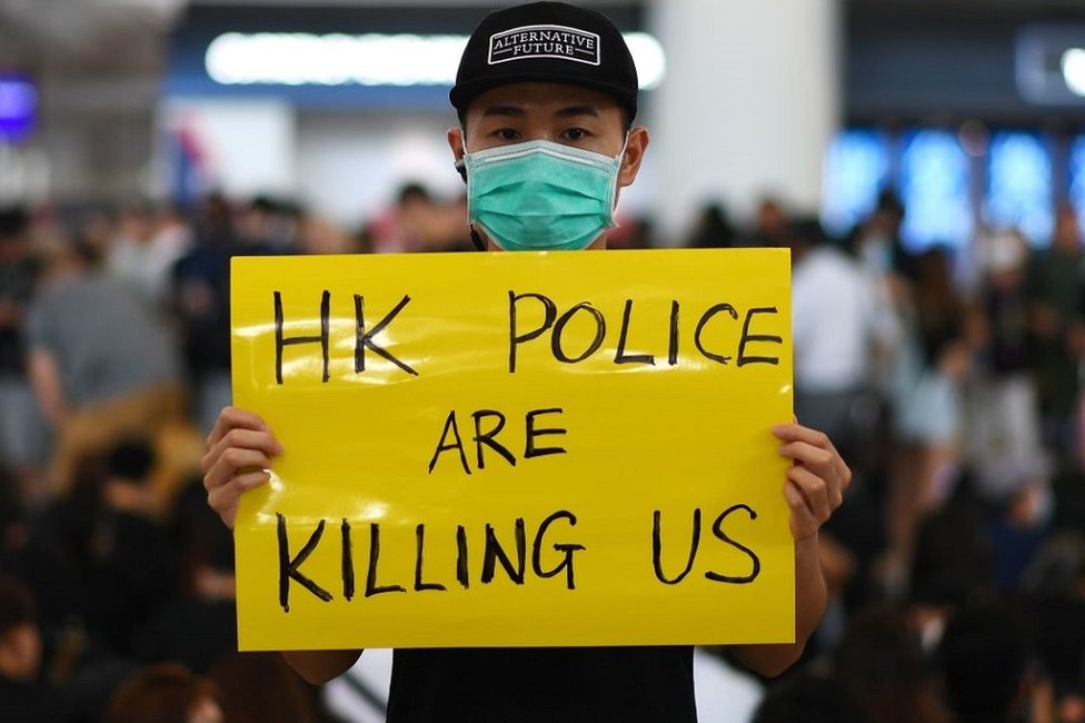 香港國際機場一號客運大樓內一名示威者舉著「香港警察在殺害我們」標語牌（12/8/2019）