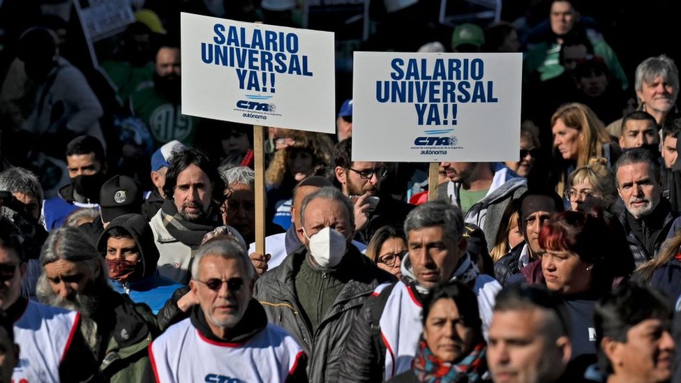 Várias pessoas protestando na rua, duas delas segurando cartaz dizendo: 'Salário universal ya!