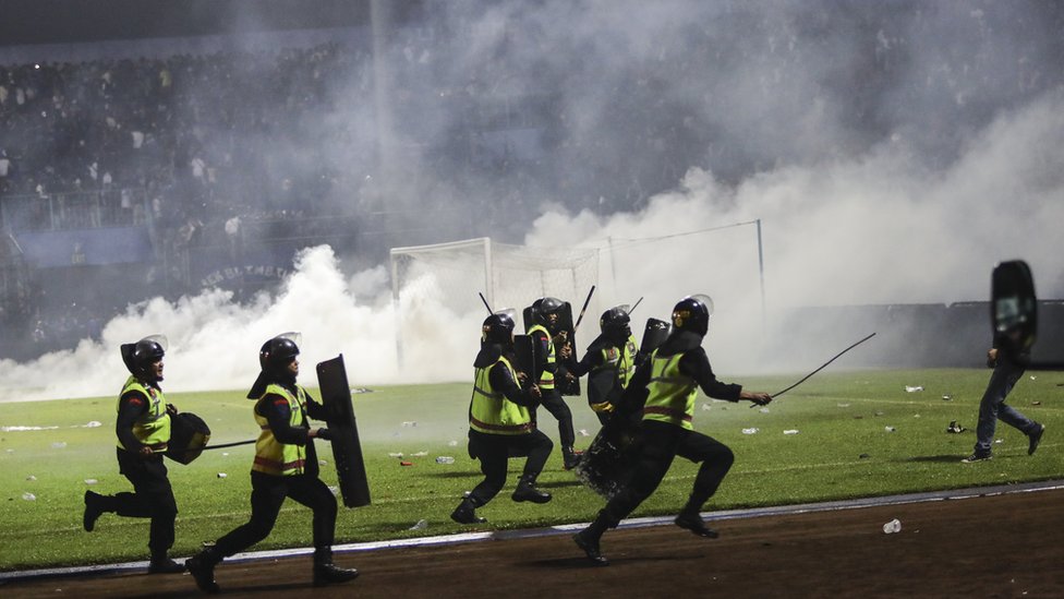 Police officers in riot gear, with shields and batons, run down the field