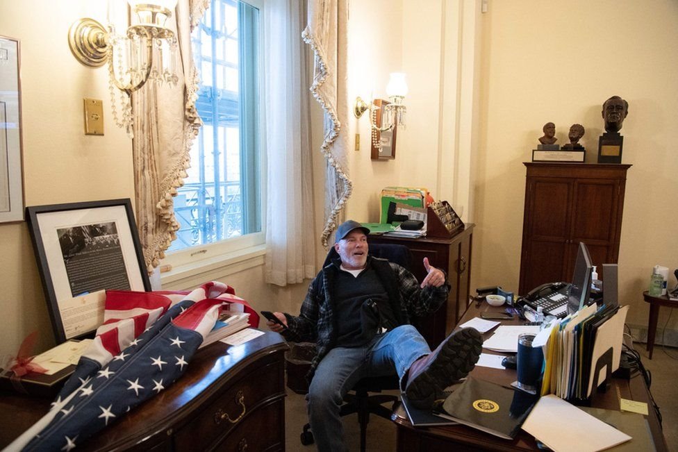 Richard Barnett, sits with one boot disrespectfully on a desk that is at the very centre of power in Congress.