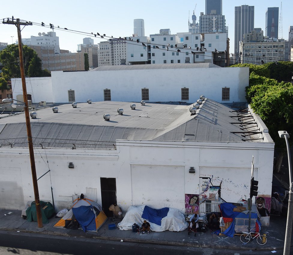 Люди проезжают на велосипедах мимо палаток на Skid Row с финансовым районом Лос-Анджелес, Калифорния, на заднем плане, 23 сентября 2015 г.