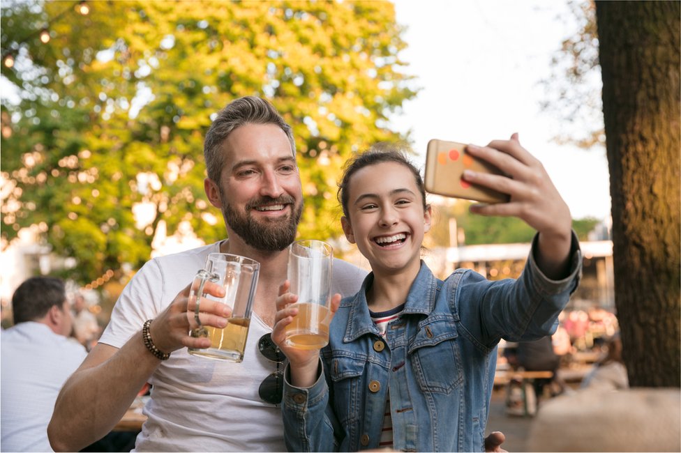 Hay padres que piensan que si sus hijos beben con ellos desarrollarán una actitud más responsable hacia el alcohol. Niñá se toma un selfie con un adulto mientras ambos sostienen un vaso de cerveza.