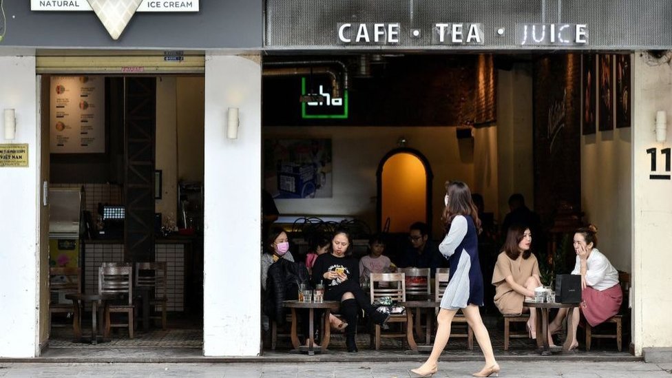 Seorang perempuan yang mengenakan masker berjalan melewati sebuah kafe tempat pelanggan duduk-duduk dan mengobrol.