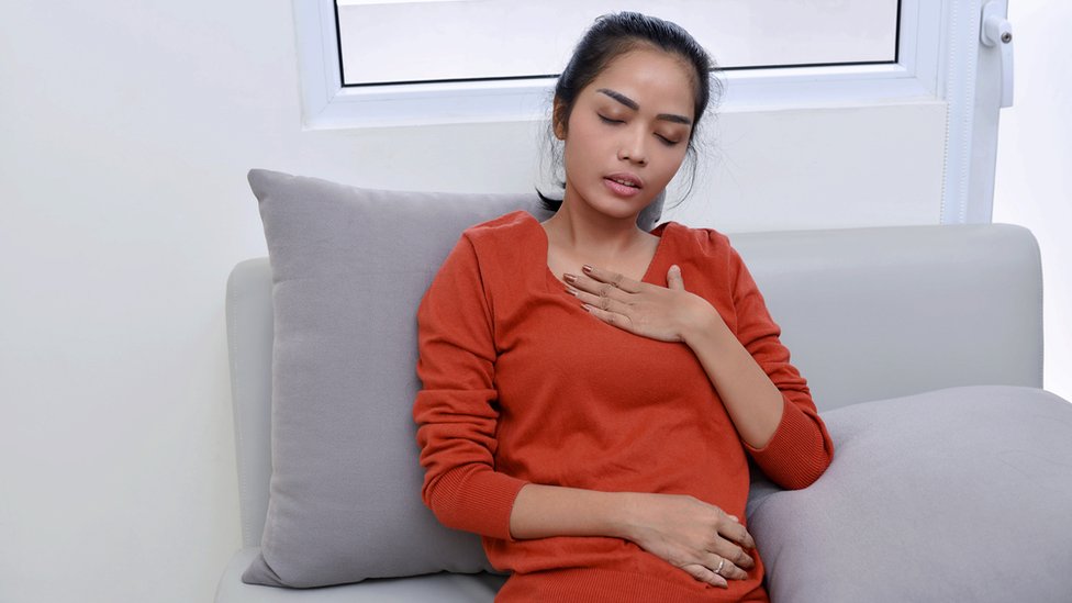 Quedarse sin aliento sin motivo puede ser síntoma de una enfermedad más grave de lo que piensas. Una mujer sentada con mano en el pecho y ojos cerrado.