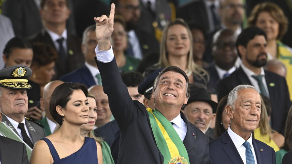 Presidente Jair Bolsonaro ao centro, com o braço apontado para o céu, acompanhado da primeira-dama e do presidente de Portugal
