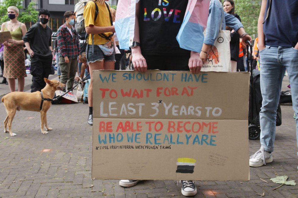 Persona sostiene cartel reclamando que no debe esperar tres años para recibir atención de afirmación de género.