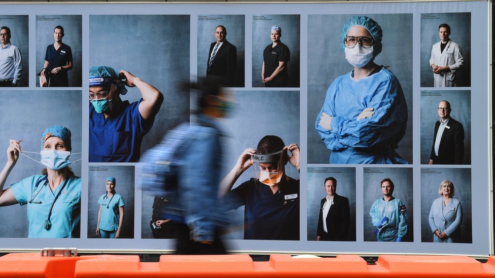 Person wearing a mask walks past a billboard in Melbourne featuring healthcare workers