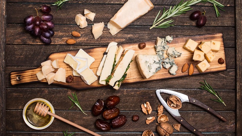 A beautifully laden cheese board, surrounded by almonds, red grapes, dates, honey, rosemary sprigs and walnuts