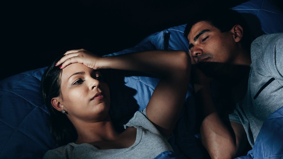 Annoyed woman lying in bed with snoring boyfriend at home in the bedroom - stock photo