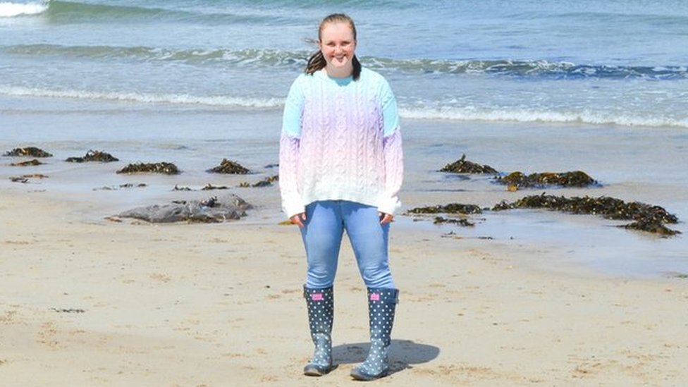 Эмма стоит на пляже с морем позади нее в радужных джинсах-джемперах и резиновых сапогах