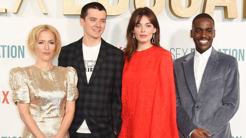 Gillian Anderson, Asa Butterfield, Emma Mackey and Ncuti Gatwa attend the World Premiere of Netflix's "Sex Education" Season 2 at The Genesis Cinema on January 8, 2020 in London. Gillian, a blonde woman in her 40s, wears a gold short sleeved dress. Asa, on her left, is a white man in his 20s with very short dark hair - he wears a checked grey and black blazer over a white T-shirt. Next to him stands Emma, a white woman in her 20s with dark hair worn loose just past her shoulders. She is wearing a long sleeved, high-necked floaty red dress. On the end, Ncuti is a black man in his 20s wearing a grey suede blazer over a white shirt and smiling at the camera.