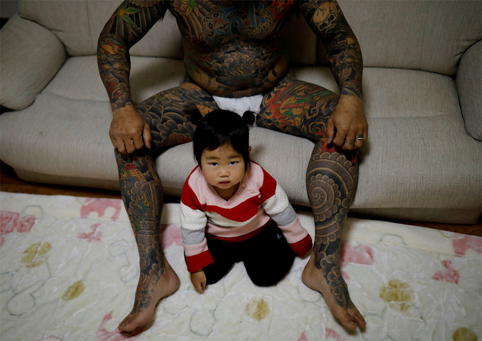 Мужчина с татуировками на ногах и туловище позирует с маленькой дочкой на ковре