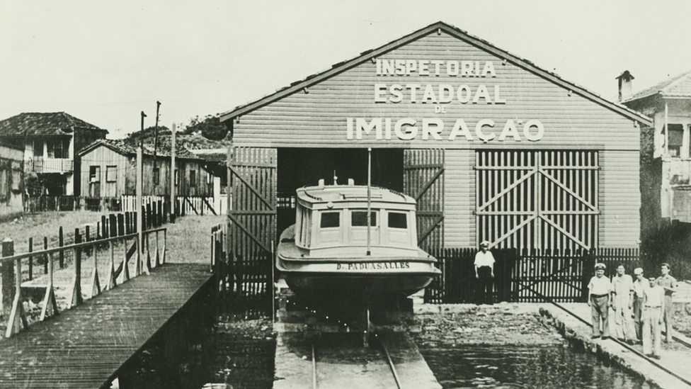 Foto em preto e branco mostra pavilhão, do qual sai barco e cuja fachada diz: Inspetoria Estadoal de Imigração