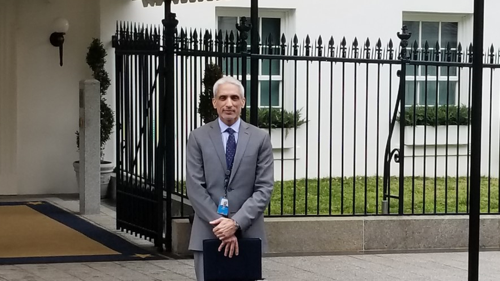 Javed Ali em pé em frente a grade da Casa Branca, segurando maleta e vestindo terno