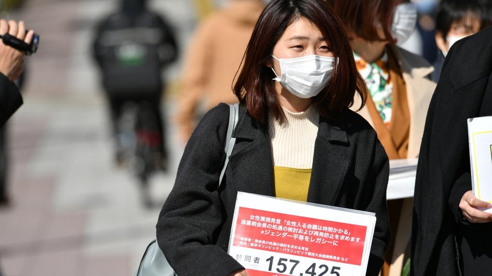 Momoko Nojo wearing a mask holds a petition with 157.425 written on it, the number of signatures collected.