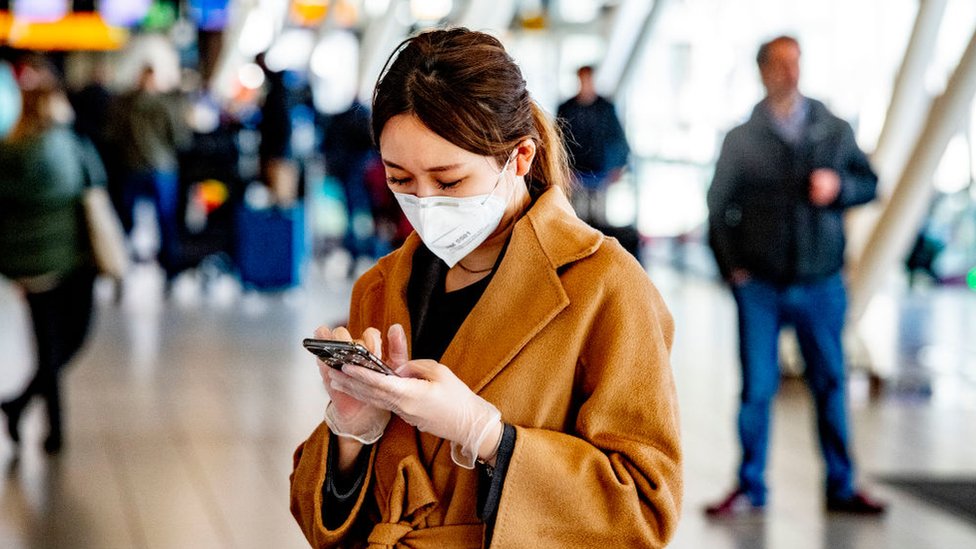 Una mujer con tapabocas y guantes sostiene un teléfono en un aeropuerto.