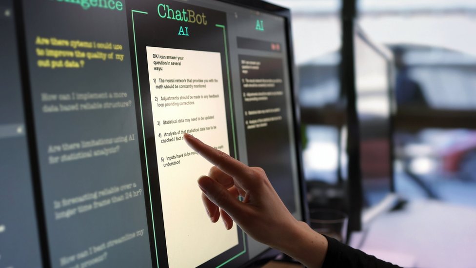 Close-up stock photograph showing a touchscreen monitor being used in an open plan office. A woman's hand is asking an AI chatbot pre-typed questions & the Artificial Intelligence website is answering.