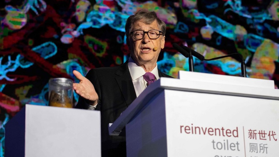 Bill Gates motions toward a jar of human faeces on stage