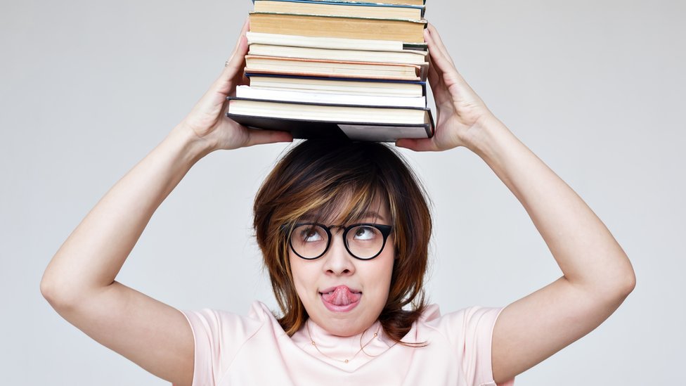 Una mujer con la lengua afuera lleva en su cabeza una pila de libros.