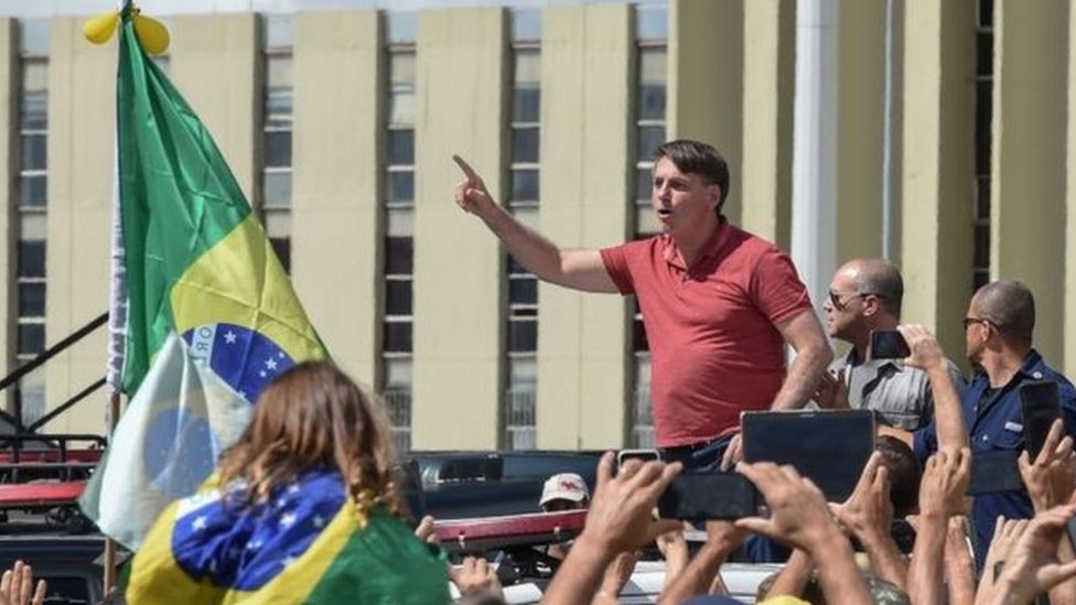 Bolsonaro em meio a manifestantes e ao lado de bandeira do Brasil, com dedo em riste