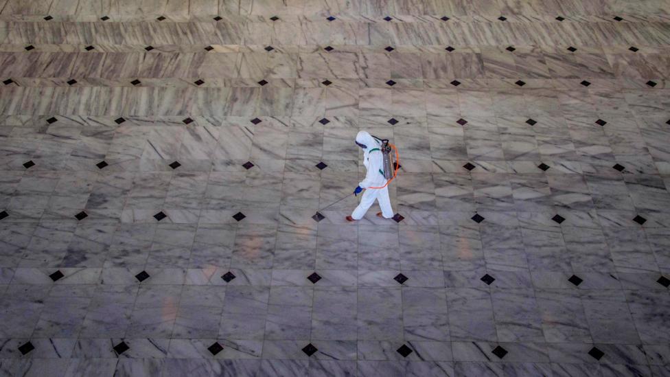 Seorang petugas kesehatan mengenakan alat pelindung diri menyemprotkan disinfektan pada lantai sebuah gedung.
