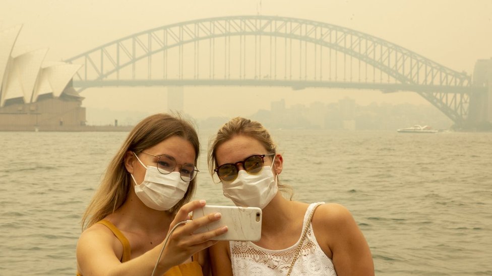 Два немецких туриста позируют для селфи в дыхательных масках, когда густой дым окутывает горизонт Сиднея