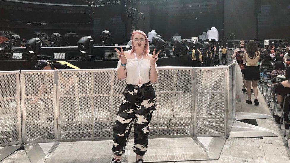 Beth is posing for a photo at a K-Pop BTS concert. She has a pink bob and is posing with her fingers held up in two v shapes. She's wearing a white top and black and white cargo pants. She is stood in front of a metal gate that is shoulder height. In the background you can see all the concert stage, stewards and lights.