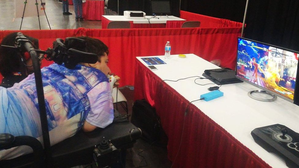 Mike playing a video game. He's lying down, flat on his stomach. A brace helps support his head, and he's operating the controller with his mouth.