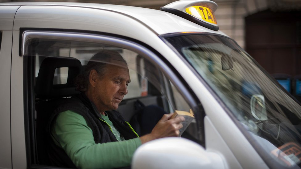Водитель такси Энди Биггс читает книгу в черном такси, стоя в очереди за проездом у вокзала Виктория в Лондоне