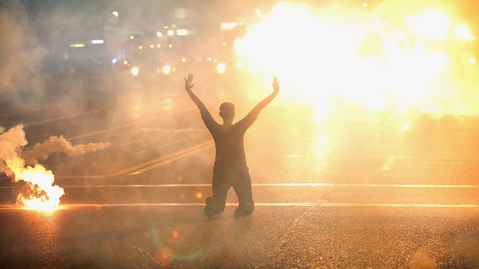 Слезоточивый газ действует на женщину, стоящую на коленях на улице с поднятыми руками после демонстрации убийства подростка Майкла Брауна сотрудником полиции Фергюсона