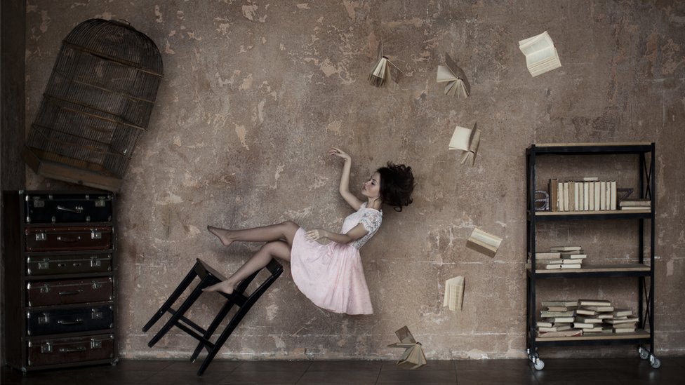 Dreamy composition of a young woman floating in a dusk pink room, with a tilted chair, flying books and a birdcage